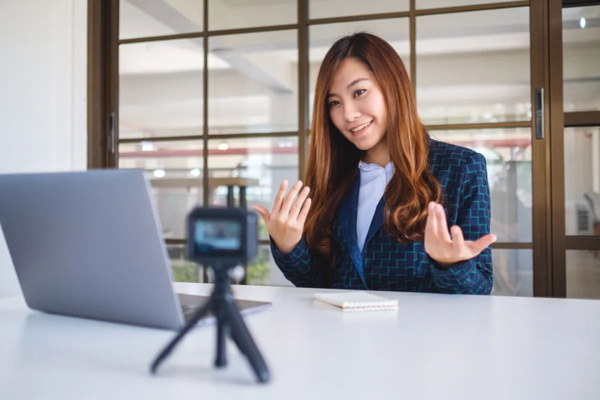 Mulher falando para uma câmera pequena posicionada diante de um notebook em uma mesa de trabalho.