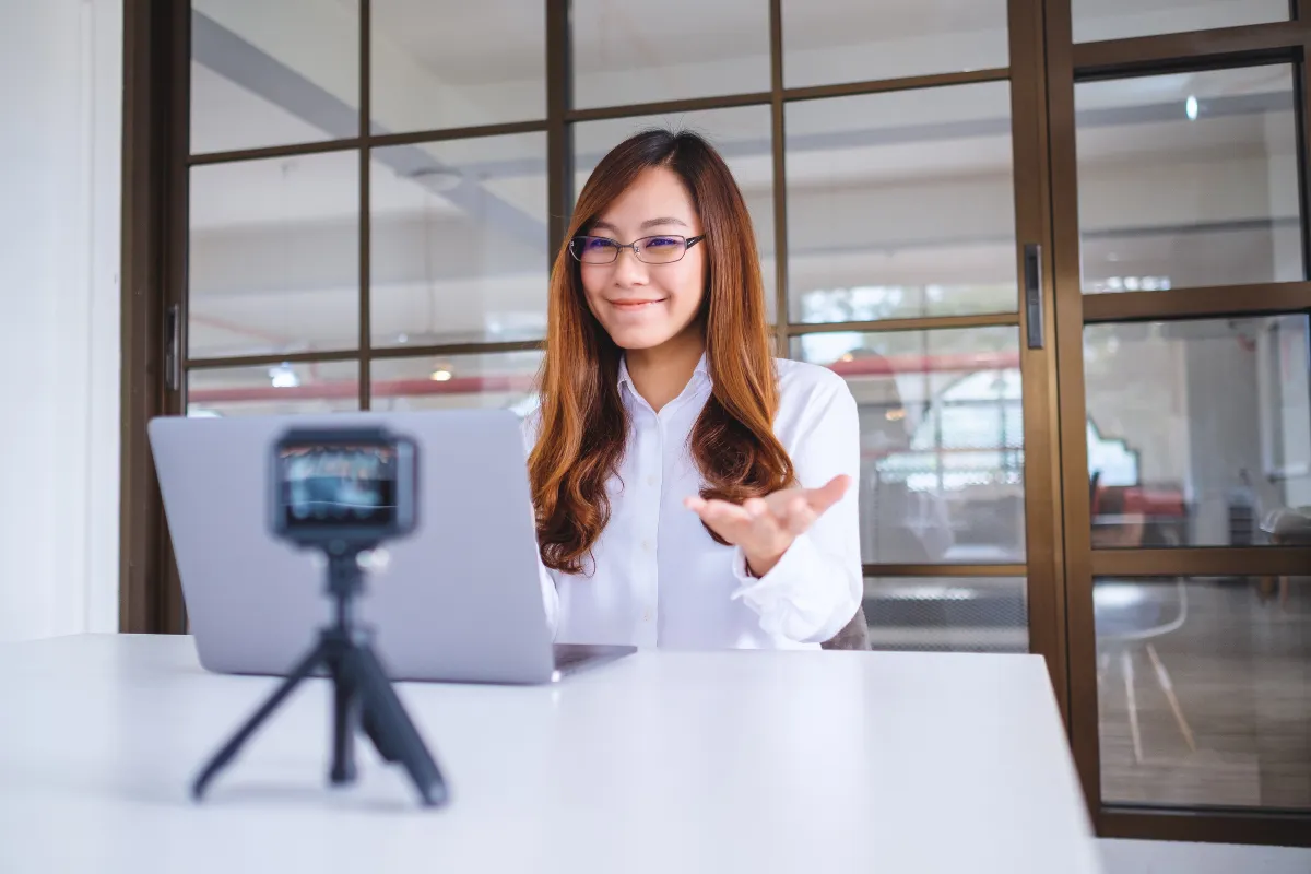 Mulher usando óculos gesticulando enquanto fala para uma câmera posicionada em frente ao notebook.