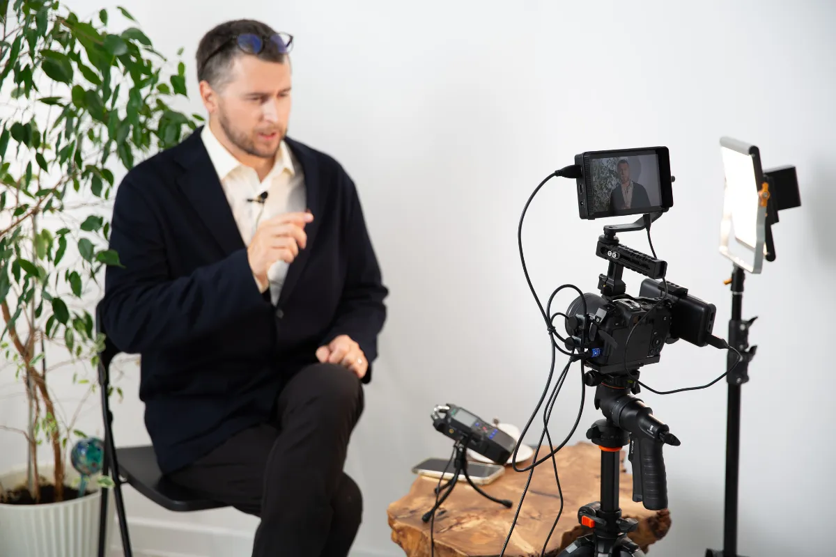 Pessoa sendo gravada por câmera com cabos visíveis, microfone e luz de apoio em ambiente interno