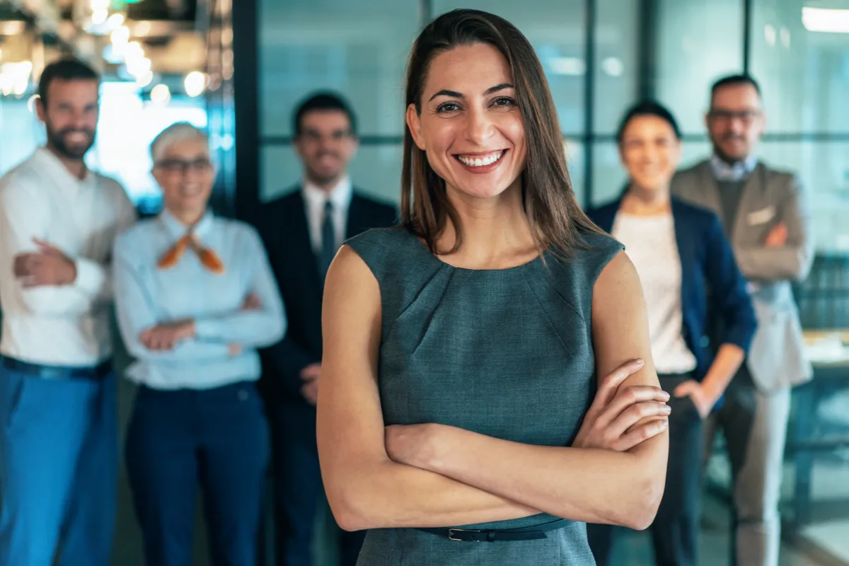 Mulher sorrindo com braços cruzados em primeiro plano com equipe desfocada ao fundo em ambiente corporativo