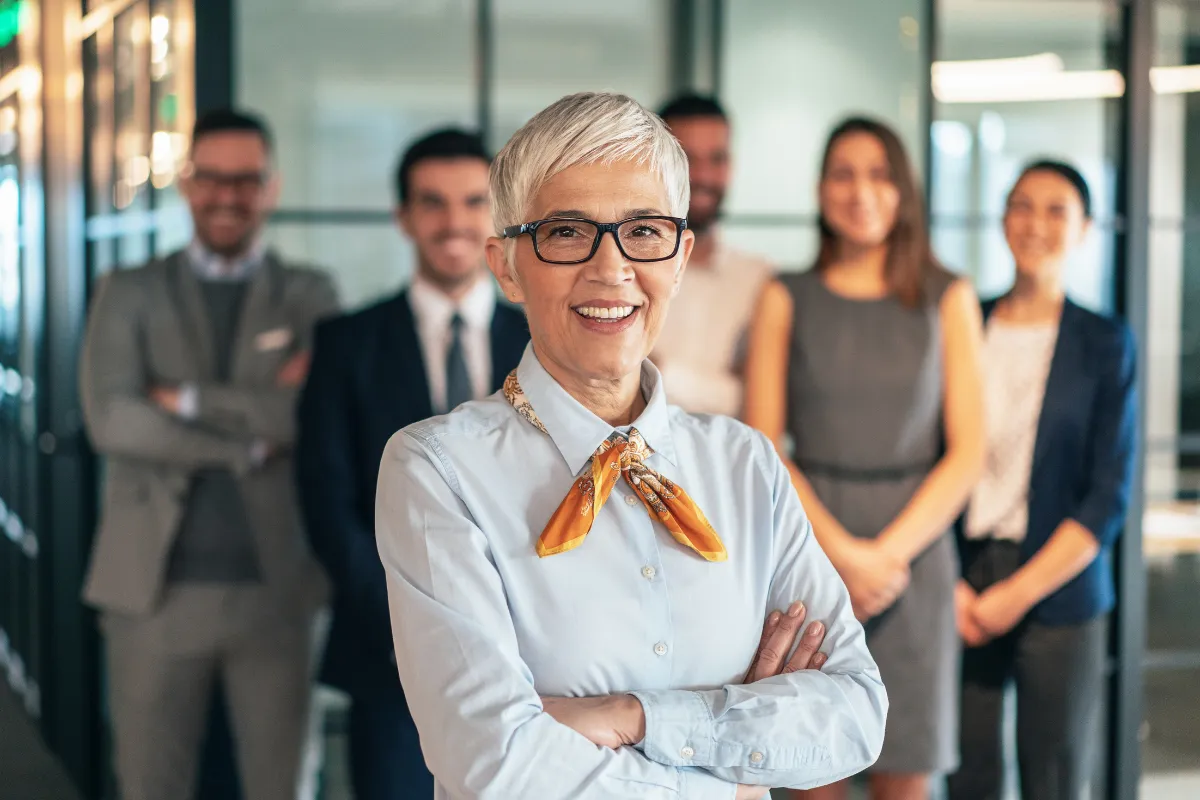 Mulher mais velha sorrindo com braços cruzados em frente a equipe de trabalho em escritório moderno